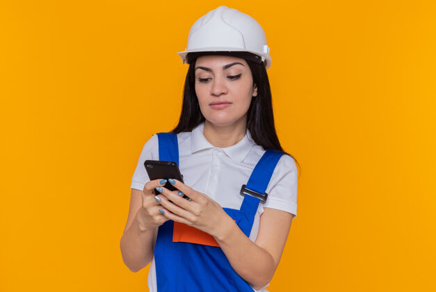留言身穿施工制服 头戴安全帽 手持智能手机 表情严肃地站在橙色墙壁上打字的年轻建筑妇女站着头盔拿着