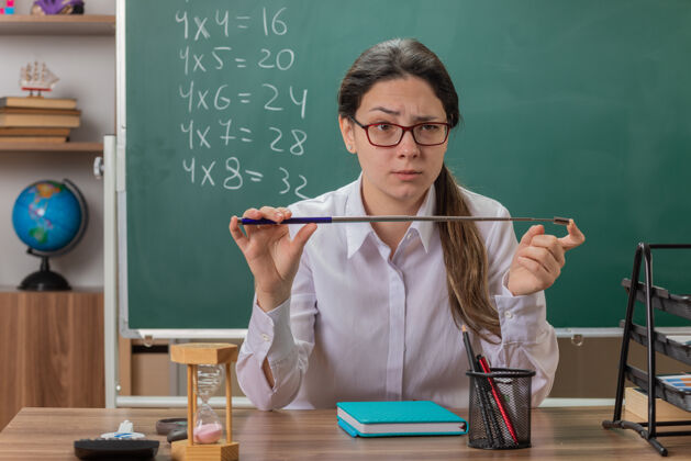拿着年轻的女老师戴着眼镜 严肃地看着前面 拿着指针 坐在教室黑板前的课桌上讲解课文脸课戴着