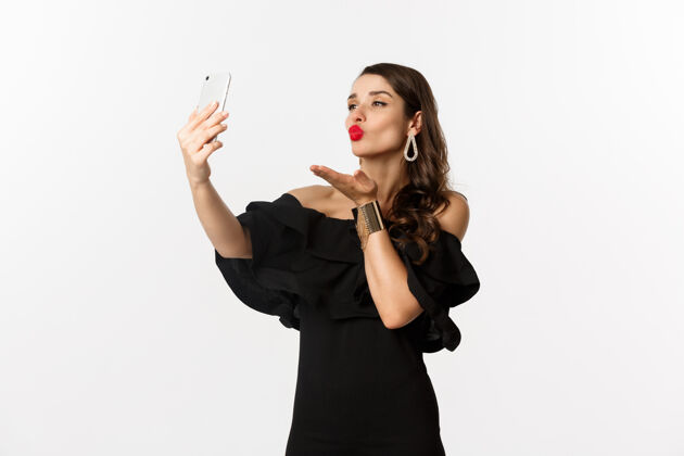 时尚穿着黑色礼服的时髦年轻女子 在手机上聚会和自拍 在智能手机摄像头前送上空中接吻 站在白色背景上莫比尔Brunette视频电话