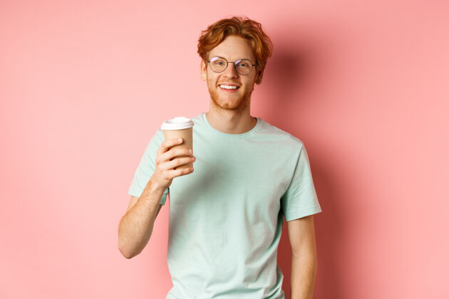 杯子戴着眼镜和t恤的快乐红发男人喝着咖啡 微笑着 享受着午休 拿着外卖杯 站在粉色的背景上咖啡学生外卖