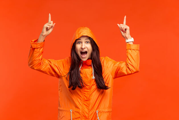 时尚在摄影棚里 一个快乐微笑的小女孩穿着秋天的橙色夹克 指着孤立的红色人类积极的情绪寒冷天气的概念雨夹克寒冷