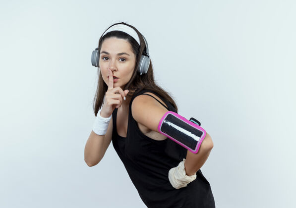 站立戴着耳机和智能手机袖标的戴着头带的年轻健身女士站在白墙上做着手指放在嘴唇上的沉默手势耳机手机品牌