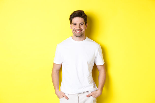 自信年轻帅哥对着镜头微笑 双手插在口袋里 站在黄色背景下工作室时尚模特