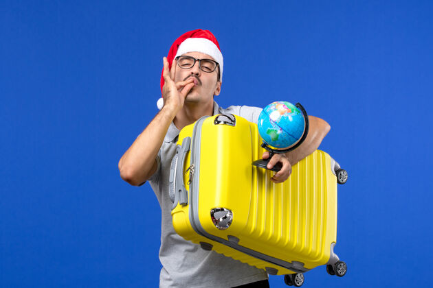运动正面图年轻男性手持地球仪和黄色袋子在蓝色墙壁飞机上度假旅行肖像圣诞节飞机