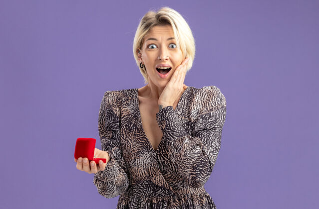 立场身着礼服的年轻美女手持红盒子和订婚戒指看着镜头幸福而惊喜的情人节概念站在紫色的墙上女人订婚期待