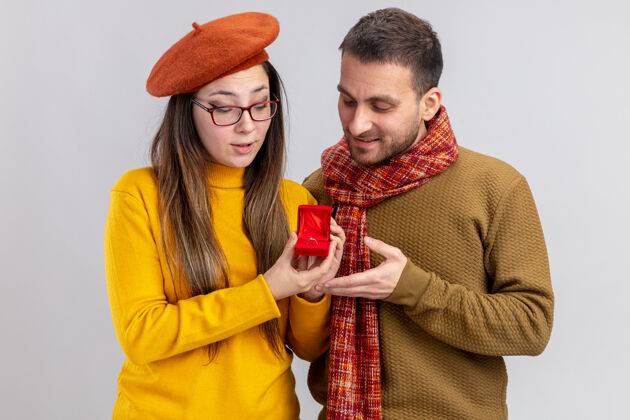 年轻年轻漂亮的夫妇幸福的男人和微笑的女人在贝雷帽与订婚戒指在红盒子庆祝情人节站在白色的背景戒指情侣订婚