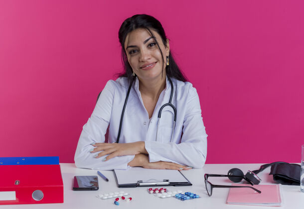 放高兴的年轻女医生穿着医用长袍和听诊器坐在办公桌旁 手放在办公桌上 隔离在粉红色的墙上工具请年轻