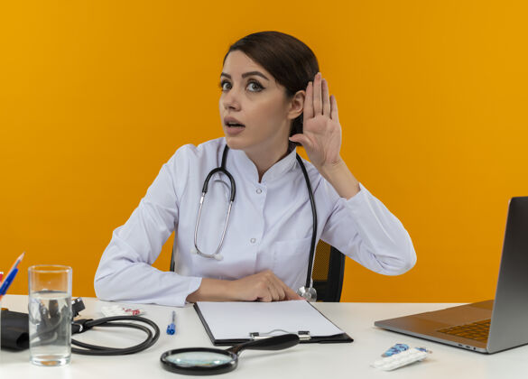 不能好奇的年轻女医生穿着医用长袍和听诊器坐在办公桌旁 拿着医疗工具和笔记本电脑直直地看着 听不到你们孤立在黄色墙上的手势年轻坐着看