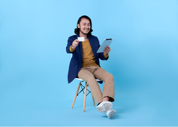 年轻亚洲人坐在明亮的蓝色椅子上 愉快地微笑着出示信用卡和使用数字平板电脑金钱互联网小工具