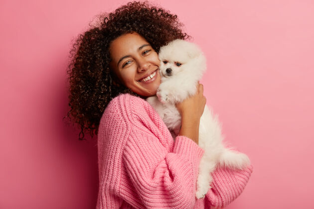 心情积极的卷发女人拥抱小狗 对宠物表达柔情 穿着针织毛衣 拜访过美容师 在粉色背景下摆姿势举行波美拉尼亚动物
