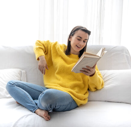 房子穿着黄色毛衣的时髦女孩正拿着一本书在家里的沙发上休息教育舒适毛衣