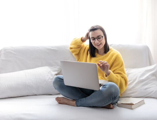女人一个穿着黄色毛衣的时尚黑发女孩坐在明亮房间的沙发上 远程操作笔记本电脑教育工作在线