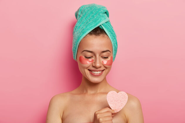牙齿可爱的欧洲女士喜欢眼部皮肤护理 把化妆海绵放在裸体上 微笑温柔 穿着裹着的浴巾 在室内做模特人与美的概念面膜黑发胶原蛋白