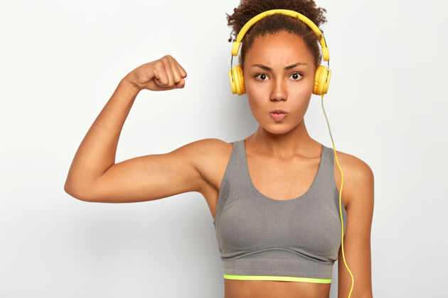 健康自信的女人表情严肃 举起手臂 展示肱二头肌 通过现代耳机听音乐 进行有氧训练人发型衣服