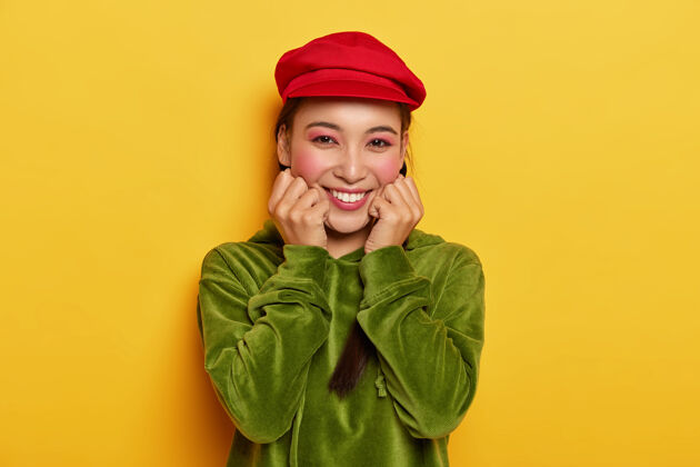 运动衫魅力柔美的东方女人 抚摸脸颊 享受美好的一天 戴着鲜红的帽子和天鹅绒绿色的运动衫衣服模仿人