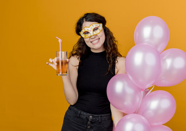 卷发年轻漂亮的卷发女子手持一束气球 戴着派对面具 快乐快乐地喝着鸡尾酒 站在橙色的墙上 生日派对的概念鸡尾酒站着女人