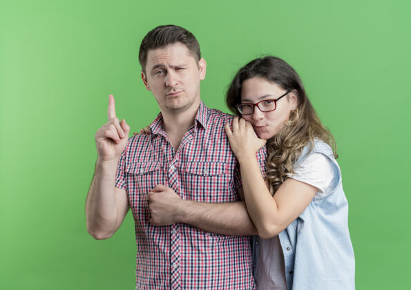 女人一对穿着休闲服的年轻夫妇站在一起 自信的男人在绿墙上展示指数斗鸡男人立场年轻人