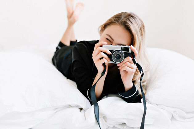 微笑快乐的年轻女模特在床上穿着睡衣 在复古相机上拍照 放松 早上在家躺在床上休闲漂亮室内