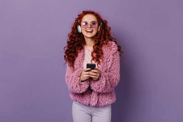 紫色背景活跃的女人戴着大耳机听音乐时会笑穿着粉色羊毛外套戴着眼镜的女孩拿着电话天后手机波浪发