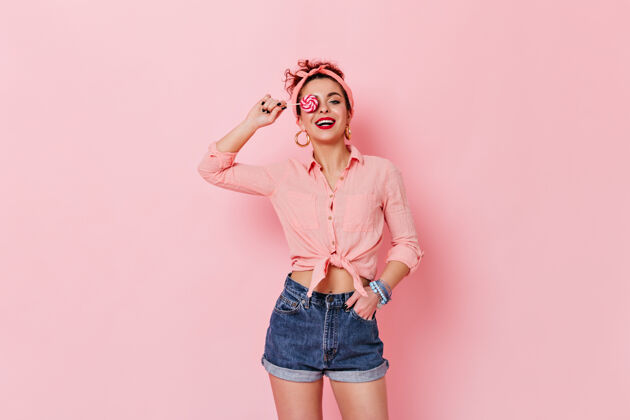 快感迷人的女人穿着别针衬衫和头带与棒棒糖在粉红色的空间摆姿势发型化妆品五十岁