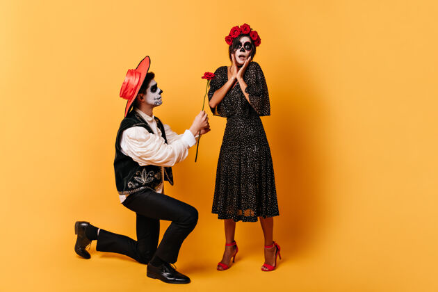年轻惊讶的戴着头盖骨面具的女人欣喜若狂 而穿着墨西哥衣服的男友跪在地上送给她红花男孩骷髅男人