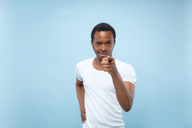 男人半身特写肖像的年轻非洲裔美国男子在白色衬衫的蓝色空间人类的情感 面部表情 广告概念指向 选择和微笑男性专业成功