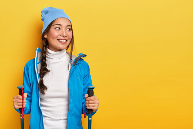 训练美丽微笑的亚洲女人喜欢斯堪的纳维亚漫步 徒步旅行 侧目 梳着辫子 戴着蓝色帽子和夹克 拿着登山竿 孤立在黄色的墙上运动客场自由空间