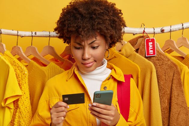女孩严肃的非洲女人用信用卡和手机在百货公司网上购物 买打折的衣服 穿着黄色的时髦衬衫 站在衣架上不同的衣服美国黄色包