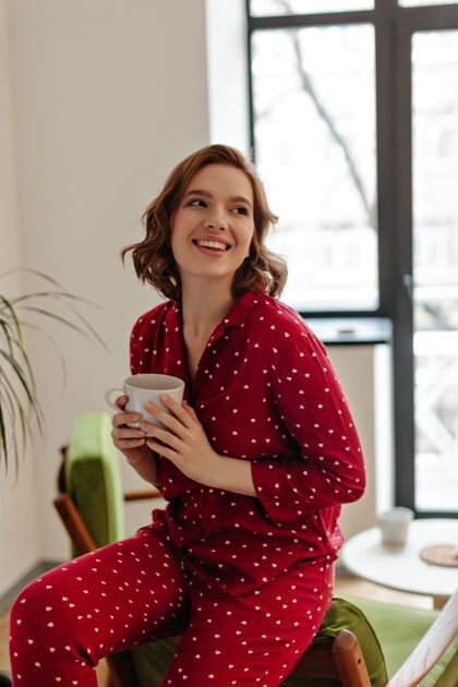 无忧无虑穿着红色睡衣的可爱女人捧着一杯咖啡室内拍摄的微笑的年轻女子在家喝茶清醒房子人