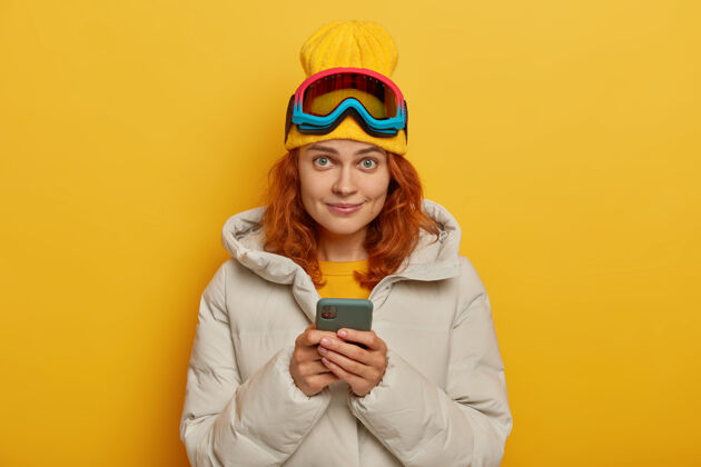 外套美丽的姜女在令人敬畏的一天后在社交网络上发布照片 在冬天积极休息 拿着手机 戴着帽子 外套和防护滑雪眼镜 在黄色的墙壁上摆姿势美国帖子非洲