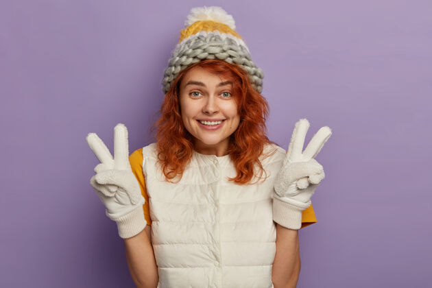 时尚喜洋洋的姜女笑容可掬 举手示意 戴着冬帽 戴着手套和白背心 心情很好 孤立在紫色的背景下白色白种人欢呼