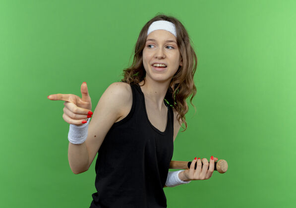 健身身穿黑色运动服的年轻健身女孩 头戴棒球棒 食指指向站在绿色墙上的一边手指立场运动装