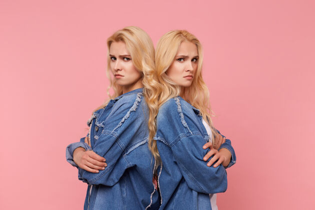悲伤不幸的年轻漂亮的长发金发姐妹们双手合十 严肃地看着镜头 背靠背地站在粉色背景下休闲外套折叠