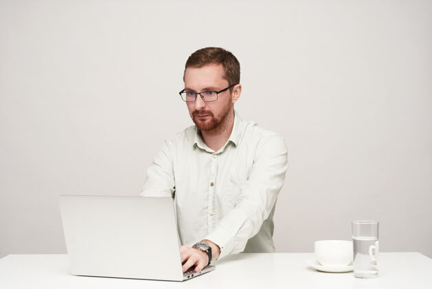 姿势专注的年轻无胡须短发男性戴着眼镜 用笔记本电脑打字 专注地看着屏幕 坐在白色背景上眼镜商务人士白色
