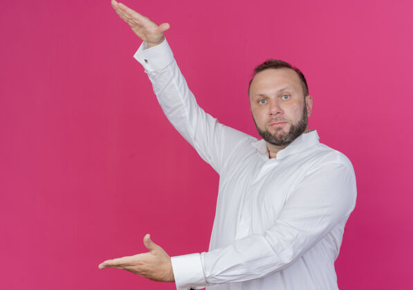胡须一个留着胡须的男人穿着白色衬衫 展示着一个大大的手势 手象征着站在粉红色的墙上穿男人手势