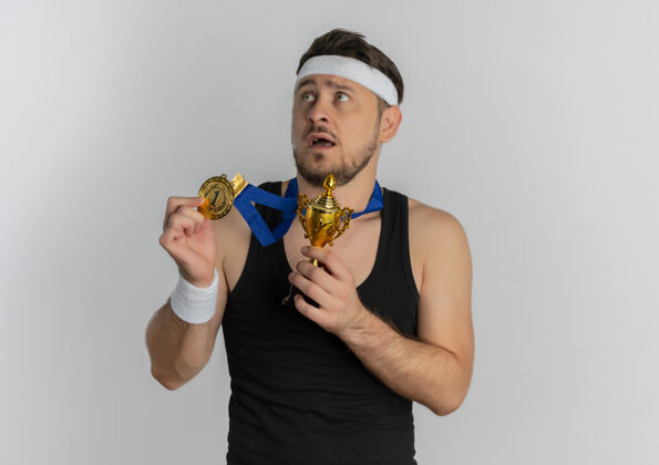 脖子戴着头带 脖子上挂着金牌的年轻健身男子拿着他的奖杯 站在白色的背景下 带着恐惧的表情望向一边周围奖牌白色