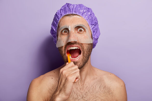 醒人物 卫生和晨间常规概念年轻人用牙刷刷牙 保持嘴巴张开 戴浴帽 脸上戴美容面具的特写镜头房子早晨白种人