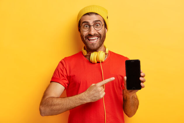 指着快乐的没刮胡子的家伙的肖像指向智能手机屏幕 演示显示器 高兴地购买新的电子设备 戴着时髦的帽子和休闲的红色t恤 模特靠着黄色的墙手指正面红色