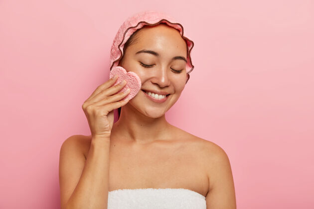 特写美丽的亚洲年轻女性的室内镜头使用化妆海绵卸妆 有问题的脂肪皮肤 集中下来与温柔的微笑 用毛巾包裹隔离在粉红色的墙壁上美丽的概念去除牙齿敏感