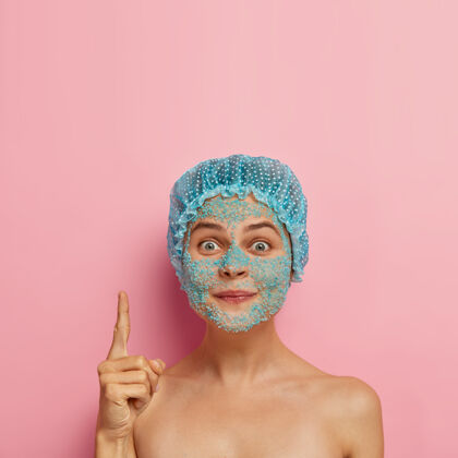 放松可爱的欧洲女人的照片 脸上有蓝色spa盐粒 头戴防水帽 食指向上指去角质姿势卫生
