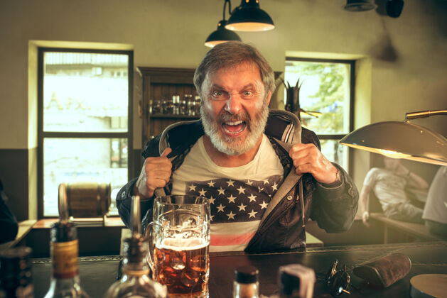 微笑大胡子男人在酒吧喝酒 看电视上的体育节目享受我最喜欢的酒和啤酒坐在桌上喝啤酒的男人足球或体育迷人类情感的概念成人啤酒饮酒