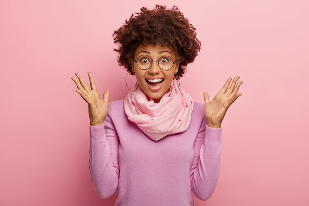 时尚可爱的快乐兴奋的女人的照片有非洲发型 提高手掌 对好消息的反应 模型在玫瑰色的工作室墙高领毛衣牙齿欣喜若狂