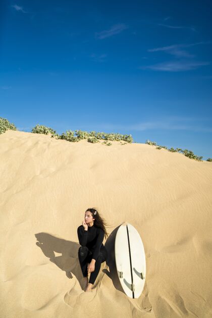 娱乐垂直拍摄一个迷人的女性坐在沙山与冲浪板在一边姿势高加索女孩