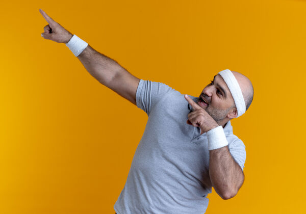 壁板戴着头巾的成熟的运动型男人看起来很自信 手指指着站在橙色墙上的一边运动站手指