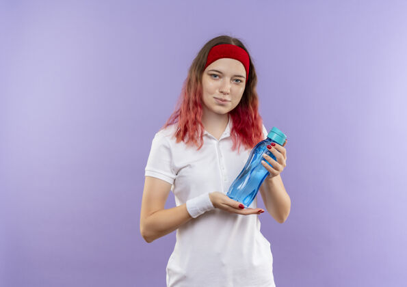 自信站在紫色的墙上 一个年轻的运动型女人拿着一瓶水 带着自信的微笑姿势水女人