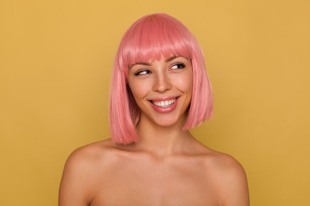 短裤年轻迷人的粉红头发女性的肖像 展示她完美的白色牙齿 一边愉快地微笑 一边站在芥末墙上积极地看着一边女性姿势鲍勃