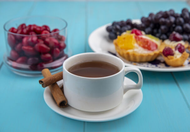 风景一杯茶的侧视图 肉桂棒和山茱萸浆果放在蓝色木制背景的玻璃碗上杯子桌子碗