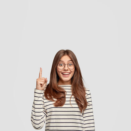 手指独立拍摄的开朗白种女人直发 展示自由空间 用食指点出你的广告内容 穿着休闲条纹毛衣 隔着白墙眼镜眼镜白种人