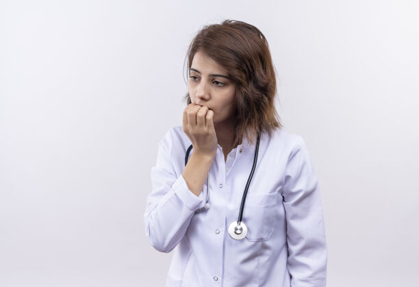 听诊器身穿白大褂 手持听诊器的年轻女医生紧张地站在白墙上咬指甲年轻人咬人医生