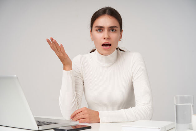 困惑激动的年轻漂亮的黑发女士 穿着白色马球衫 困惑地看着镜头 困惑地举起她的手掌 坐在桌旁 手里拿着现代化的笔记本电脑衣服站立笔记本电脑